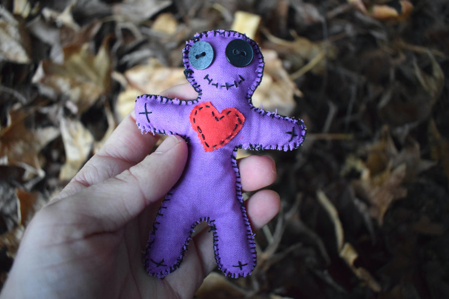 A hand holding a purple hoodoo voodoo dolls with black button eyes, a stitched smile and red fabric love heart on its heart nestled on a bed of leaves.