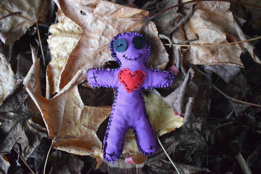 A purple fabric hoodoo voodoo doll with black button eyes, a stitched smile and red fabric love heart on its heart nestled on a bed of leaves.