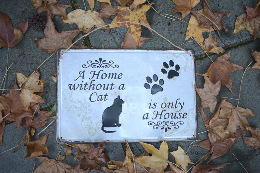 A metal sign with a black cat and two black paw prints saying A Home without a Cat is only a House on it resting on a bed of leaves