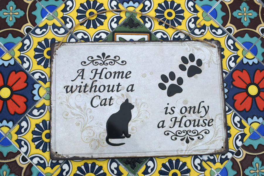 A metal sign with a black cat and two black paw prints saying A Home without a Cat is only a House on it resting on a mosaic background