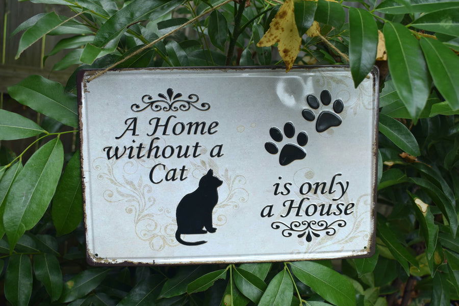 A metal sign with a black cat and two black paw prints saying A Home without a Cat is only a House on it nestled against greenery