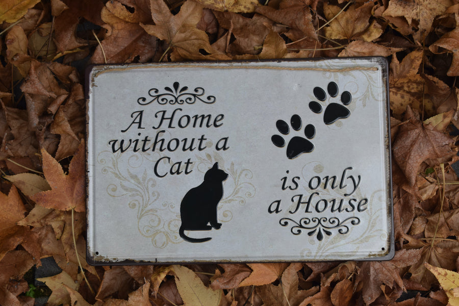 A metal sign with a black cat and two black paw prints saying A Home without a Cat is only a House on it resting on a bed of leaves