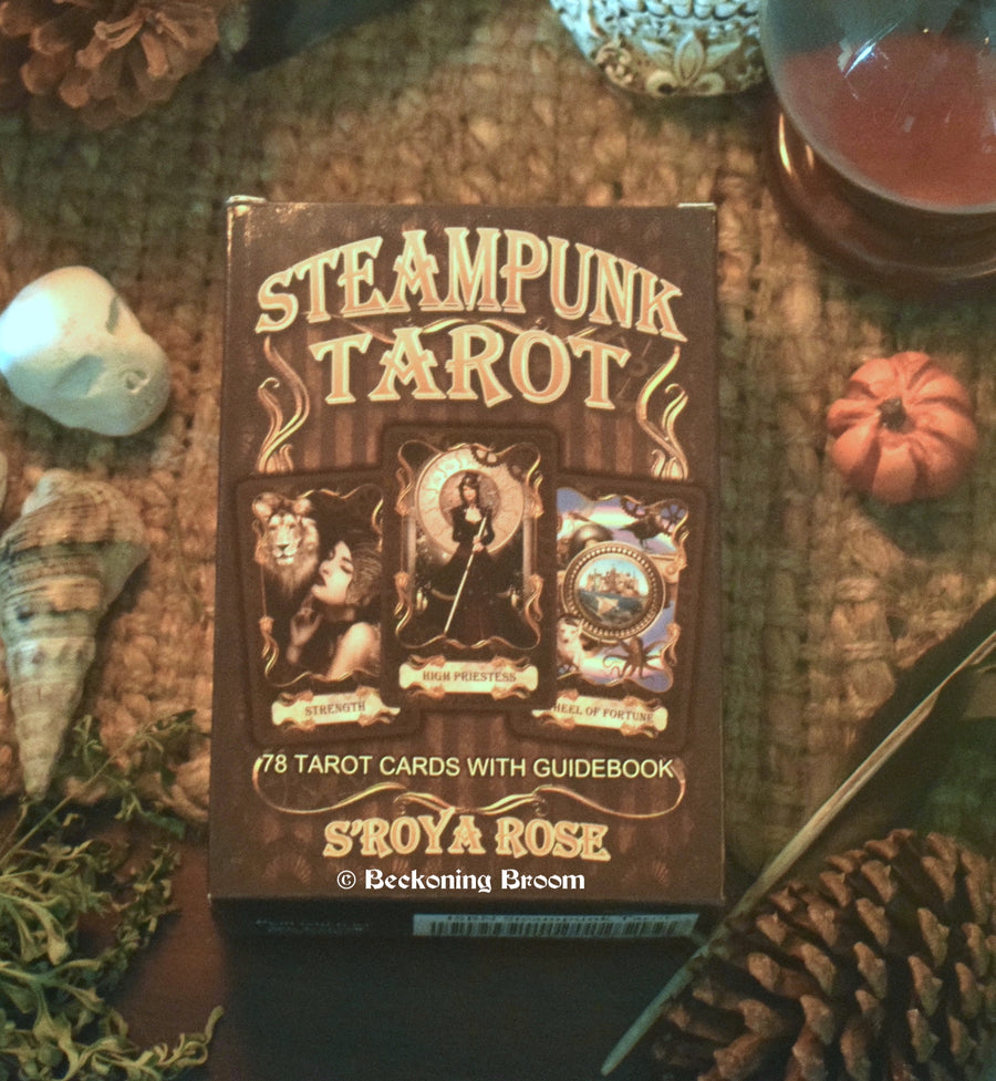A tarot deck with Steampunk Tarot S'roya Rose written on the front rests on an altar with a crystal ball, shell, herbs and other mysterious objects.