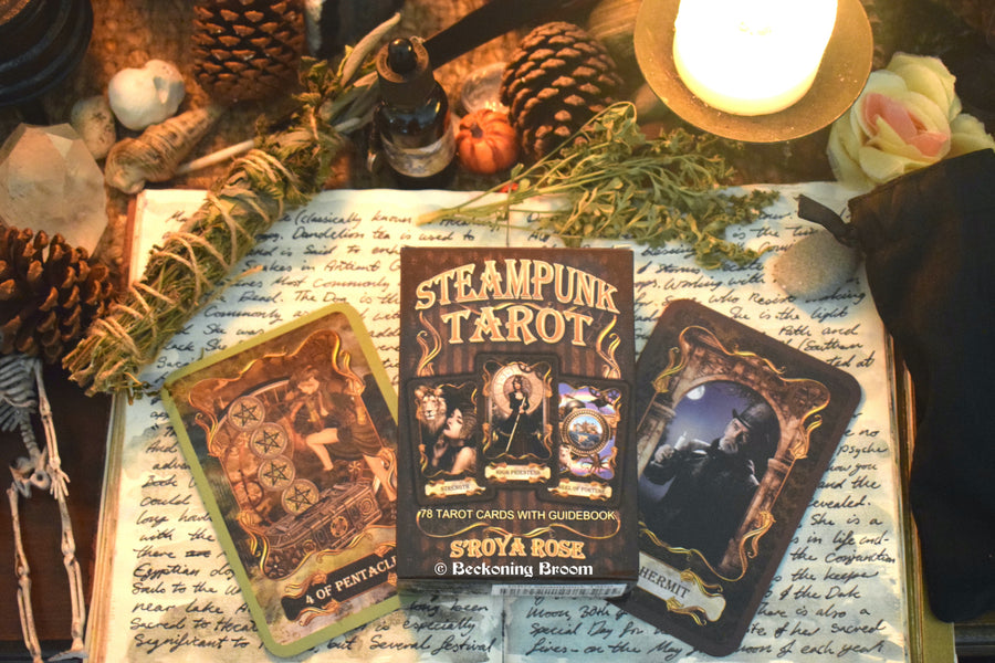 A tarot deck with Steampunk Tarot S'roya Rose written on the front with two cards laid out on an open grimoire with candles, herbs and other mysterious items surrounding.