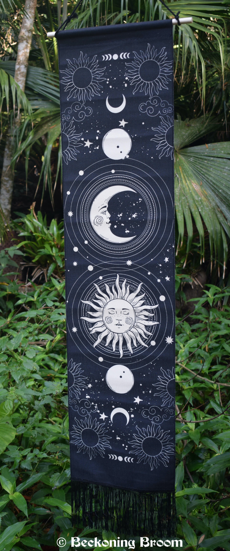 A black fabric tapestry depicting white moons and a sun with a long black fringe hangs with greenery in the background.