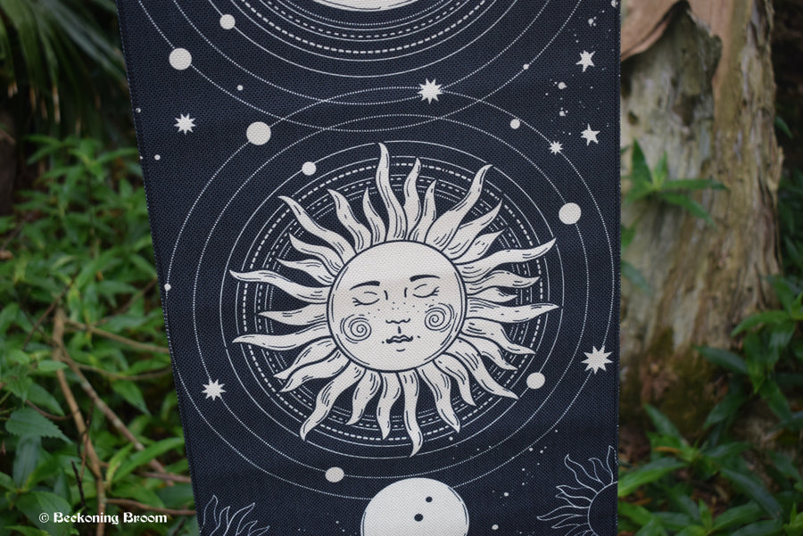 A detail of a black fabric tapestry depicting a white sun hangs with greenery in the background.