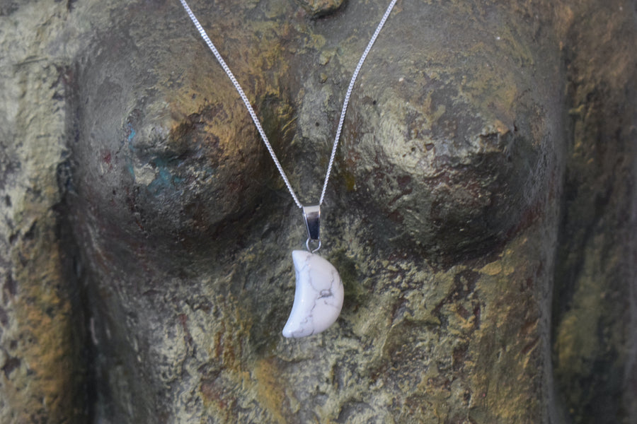 A white howlite crystal crescent moon necklace on a sterling silver chain hanging on the neck of a bronze goddess sculpture