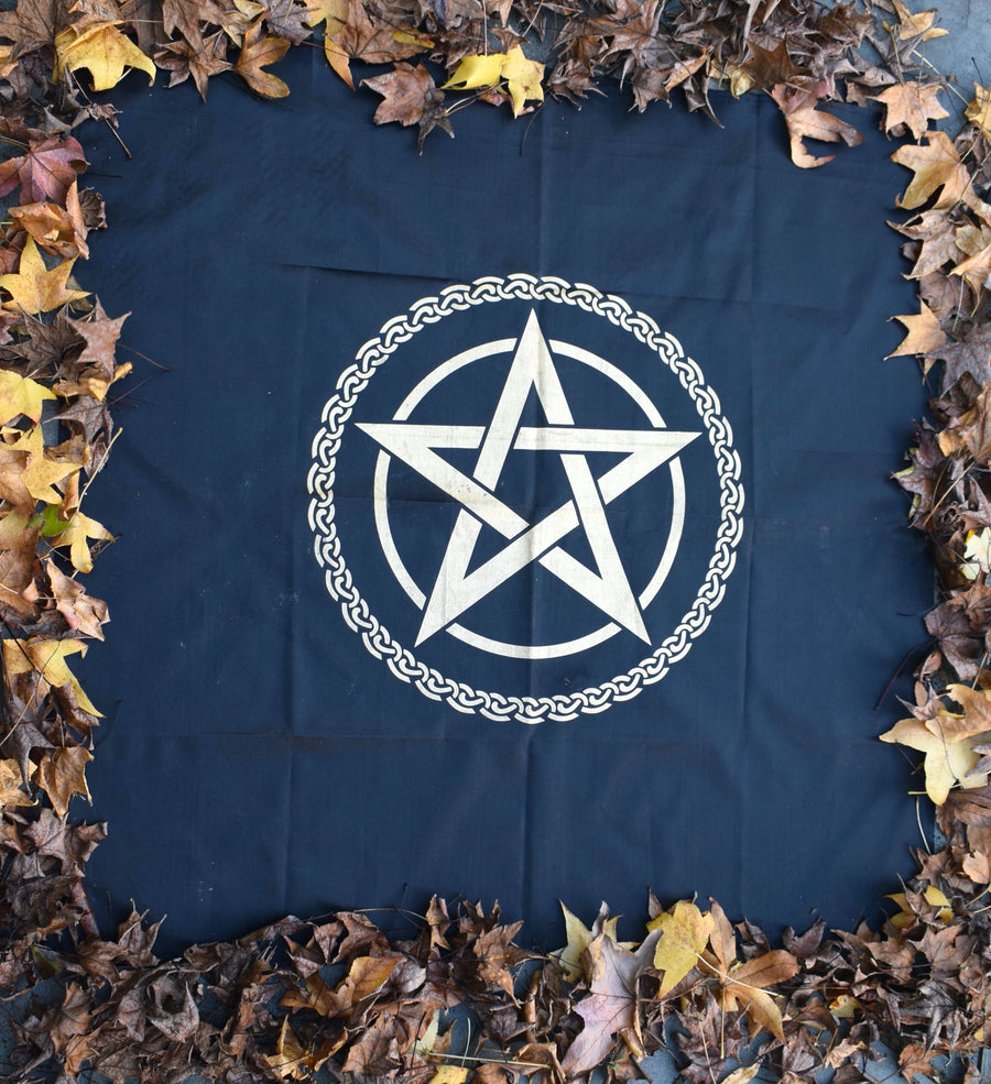 A black satin altar cloth or witches flag with gold pentagram pentacle in the middle encircled by a celtic knot on a bed of autumn leaves