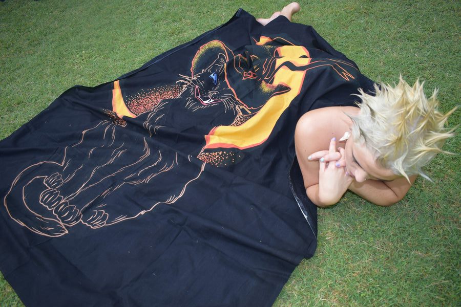 Person lying on grass with a black hand painted fabric throw depicting a black cat wearing witches hat outlined in yellow and orange with yellow crescent moon in background draped over them