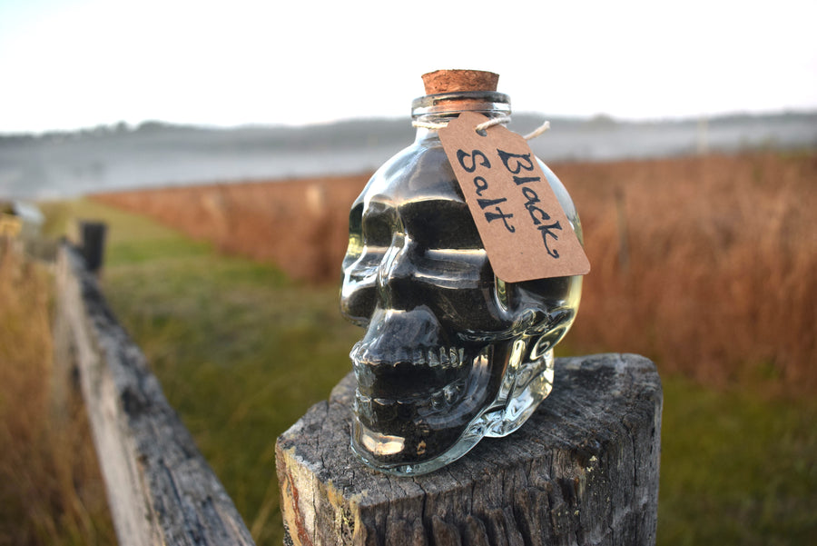 Glass skull jar filled with black salt, ground witches burrs and yule ash sitting on an old fence paling with grass, mist and mountains in the background 