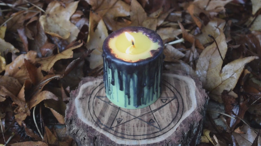 A green pillar candle with black drips sits with a flickering wick alight on a pentagram wooden disk nestled on a bed of leaves.