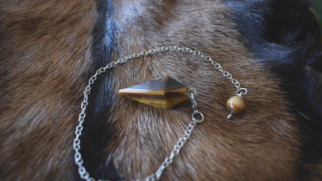 Small Tiger's Eye point crystal pendulum with chain and bead resting on goatskin fur and hand holding undulating pendulum