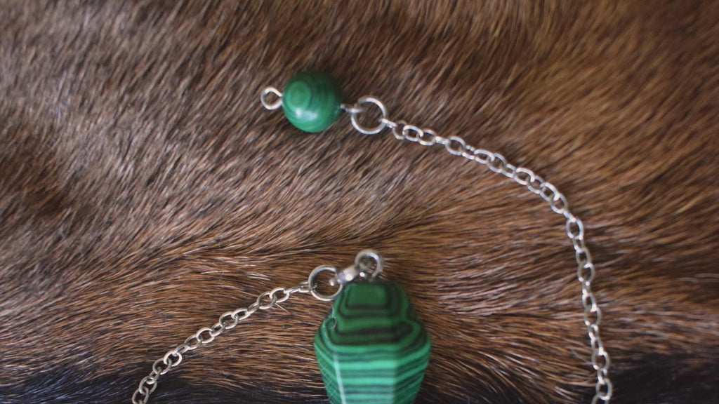 Small malachite point crystal pendulum with chain and bead resting on goat skin fur and undulating while being held