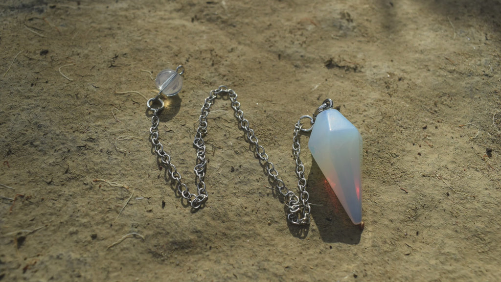 Small opalite point crystal pendulum with chain swinging in the air with trees in background