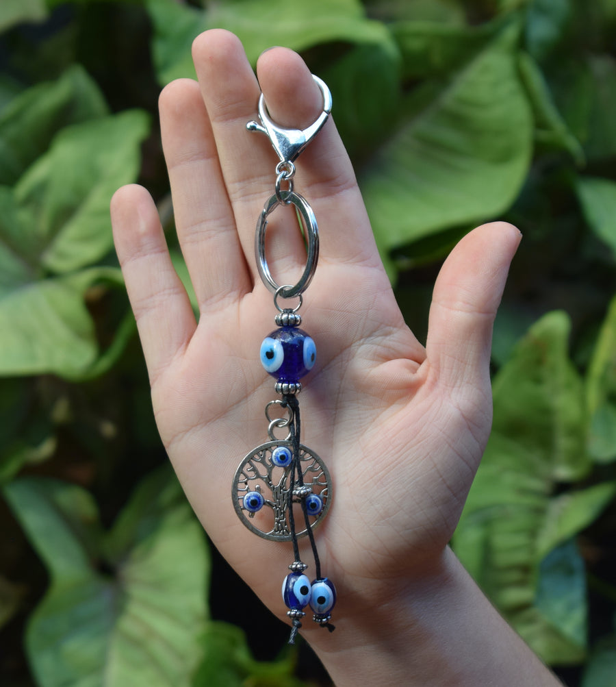 A hand holds an evil eye tree of life key ring with greenery in the background.