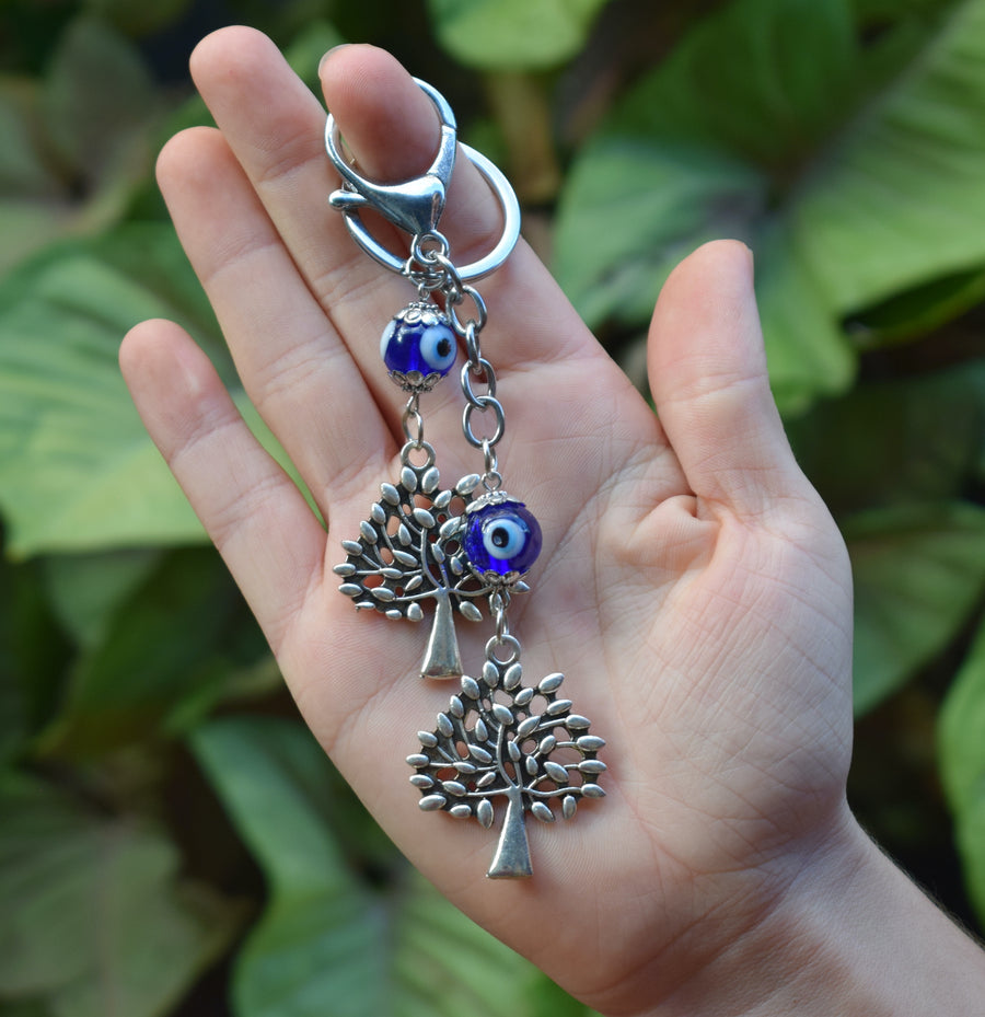 A hand holds a key ring with two evil eye beads and two silver coloured leafy trees with greenery in the background.