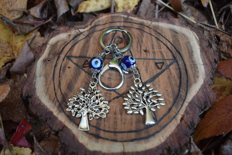 A key ring with two evil eye beads and two silver coloured leafy trees rests on a wooden pentagram disk nestled on autumn leaves.