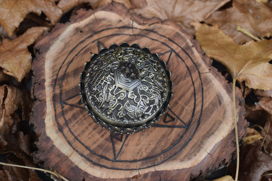 A metal lotus trinket box with a lid resting on a wooden pentagram trivet nestled on a bed of autumn leaves