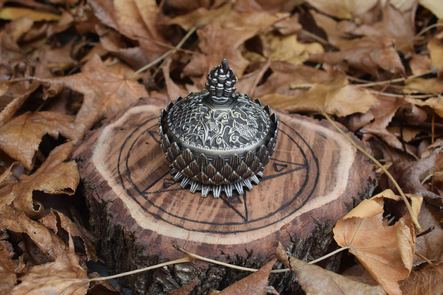 A metal lotus trinket box with a lid resting on a wooden pentagram trivet nestled on a bed of autumn leaves