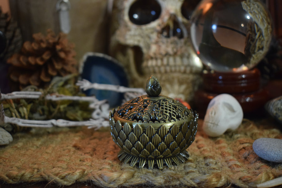 A metal lotus trinket box with a lid resting on a altar with skulls, herbs and a crystal ball in the background