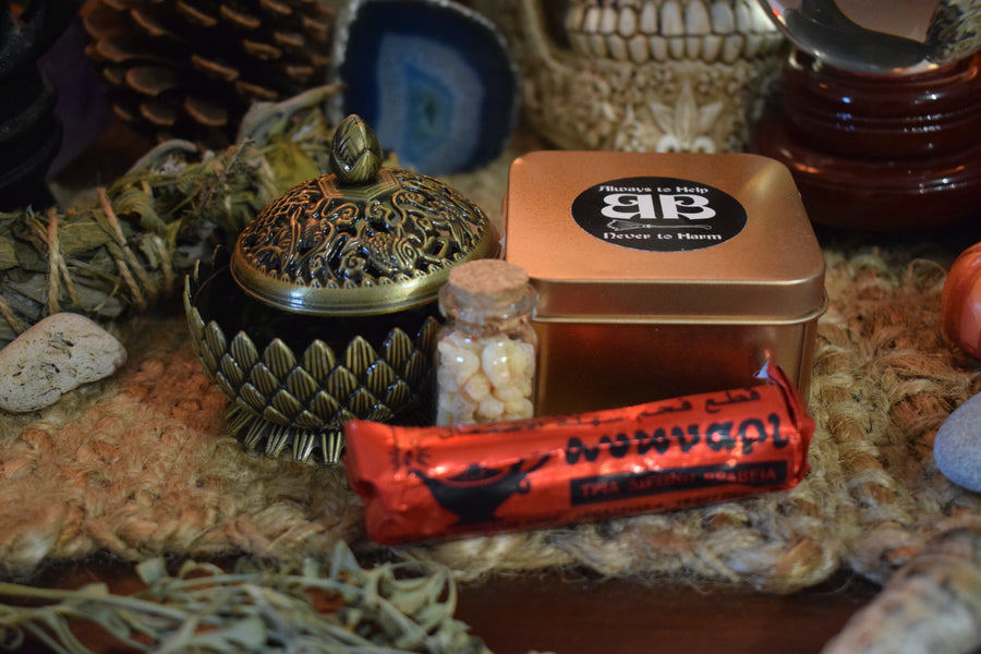 A metal lotus trinket box with a lid resting on an altar with frankincense resin, charcoal disks, herbs and crystals in the background