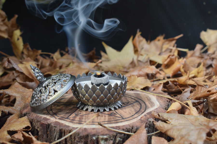 A metal lotus trinket box with a lid half off with smoke wafting up into the air resting on a wooden pentagram trivet nestled on a bed of autumn leaves