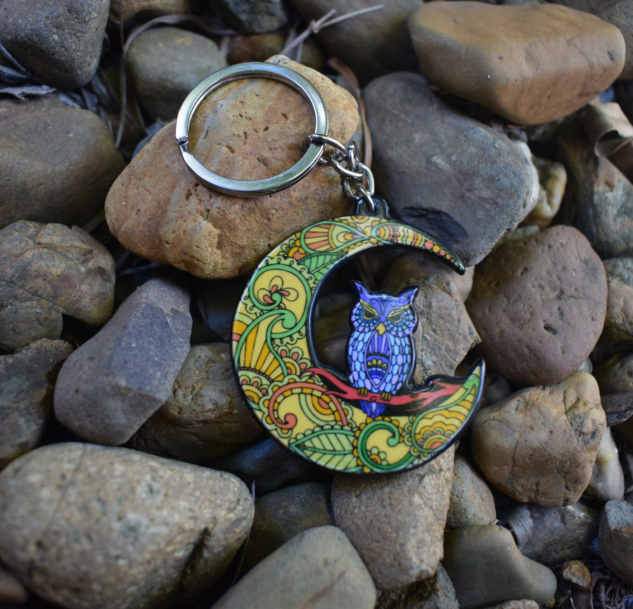 A key ring with a resin blue owl perched on a colourful crescent moon on a rocky background.