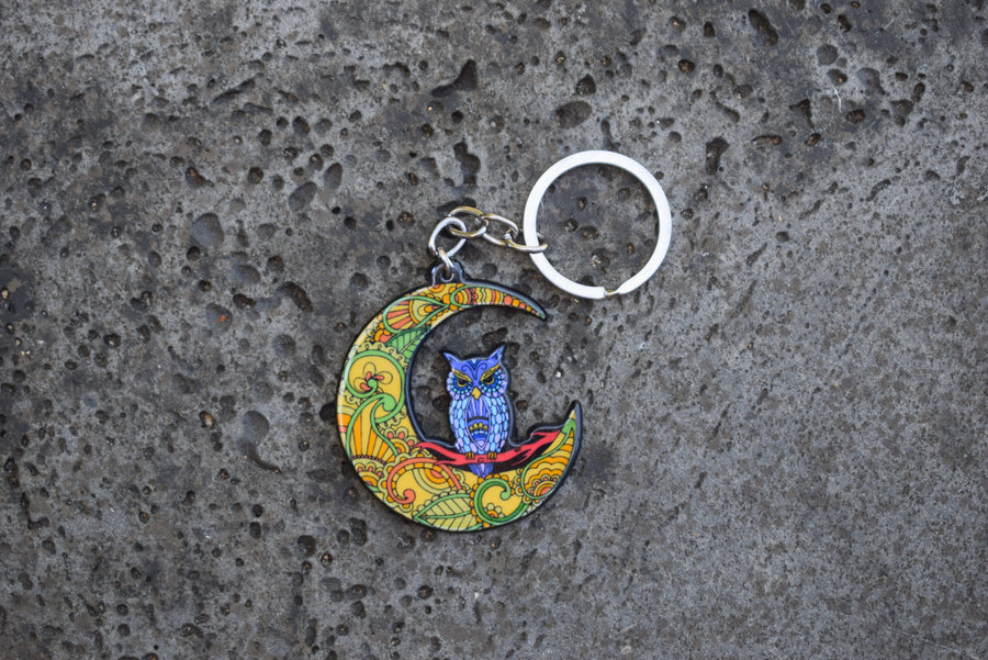 A key ring with a resin blue owl perched on a colourful crescent moon on a rock background.