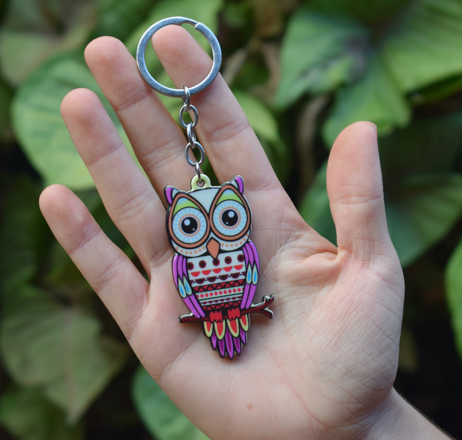 A hand holds a key ring in the shape of an owl sitting on a branch with greenery in the background.