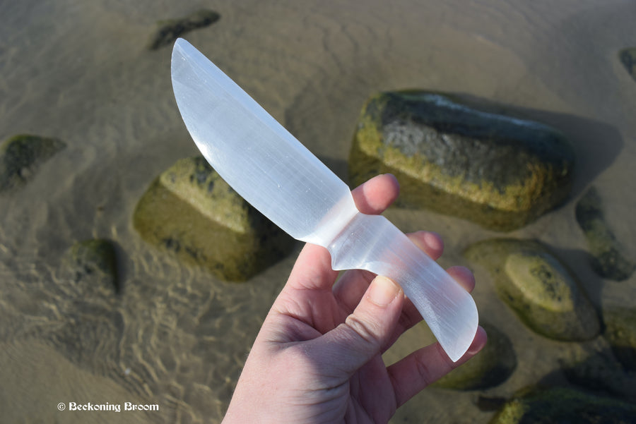 A hand holding a selenite ceremonial blade with rocks and water in the background.