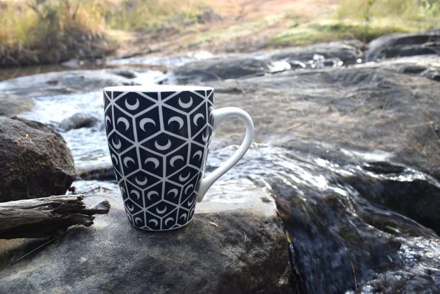 A black and white cup with crescent moon and sacred geometry design resting on a rock near a stream