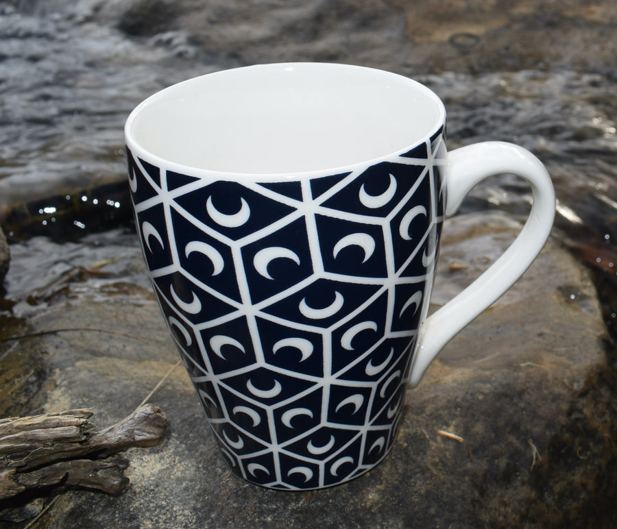 A black and white cup with crescent moon and sacred geometry design resting on a rock near a waterfall