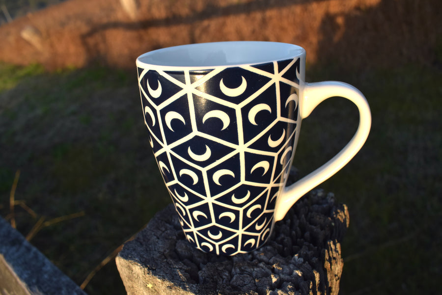 A black and white cup with crescent moon and sacred geometry design resting on fence palings