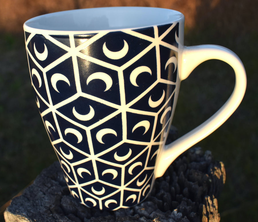 A black and white cup with crescent moon and sacred geometry design resting on fence palings