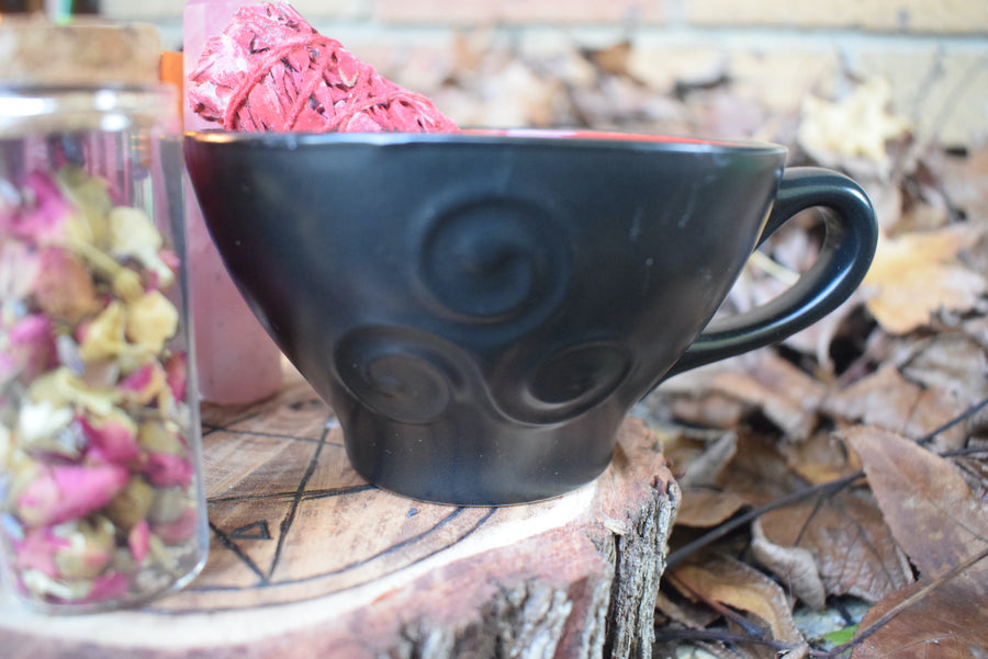 A black tea cup with a triple moon goddess symbol on it sits on a piece of wood with a pentagram on it with herbs and a smudge stick