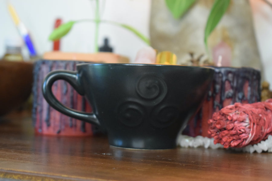 A black tea cup with a triple moon goddess symbol on it sits with herbs and a smudge stick