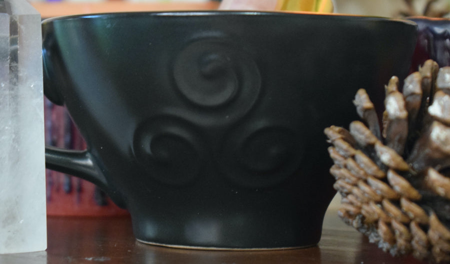 A black tea cup with a triple moon goddess symbol on it sits with herbs and a smudge stick
