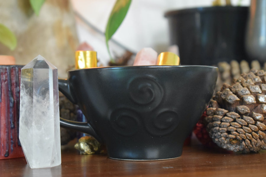 A black tea cup with a triple moon goddess symbol on it sits with crystal and pinecone