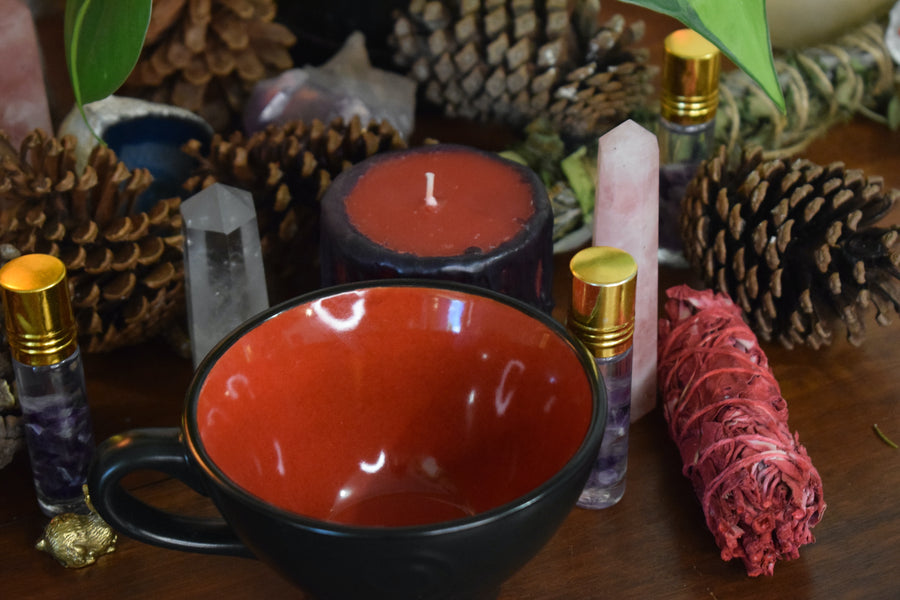 A black tea cup with a triple moon goddess symbol on it and red inside sits on an altar with herbs, crystals, pinecones and a smudge stick