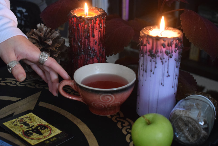 A hand reaches for a pink orange tea cup with a triple moon goddess symbol on surrounded by candles, tarot cards and snake skin