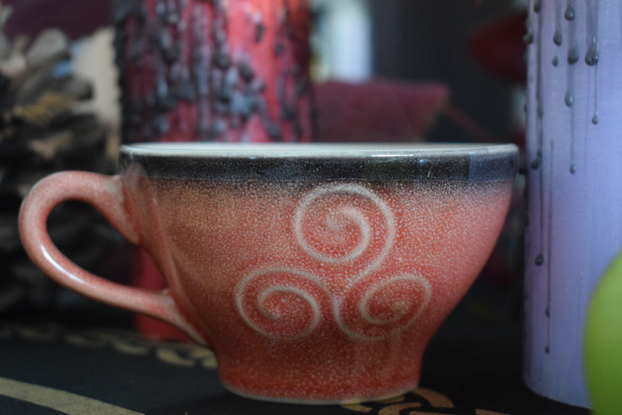 A pink orange tea cup with a triple moon goddess symbol on surrounded by candles