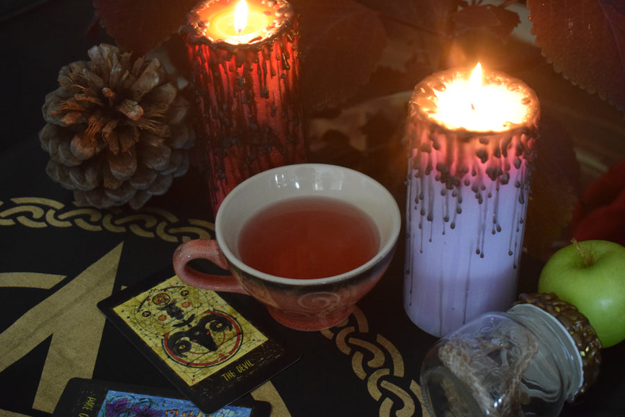 A pink orange tea cup with a triple moon goddess symbol on surrounded by candles, tarot cards and snake skin