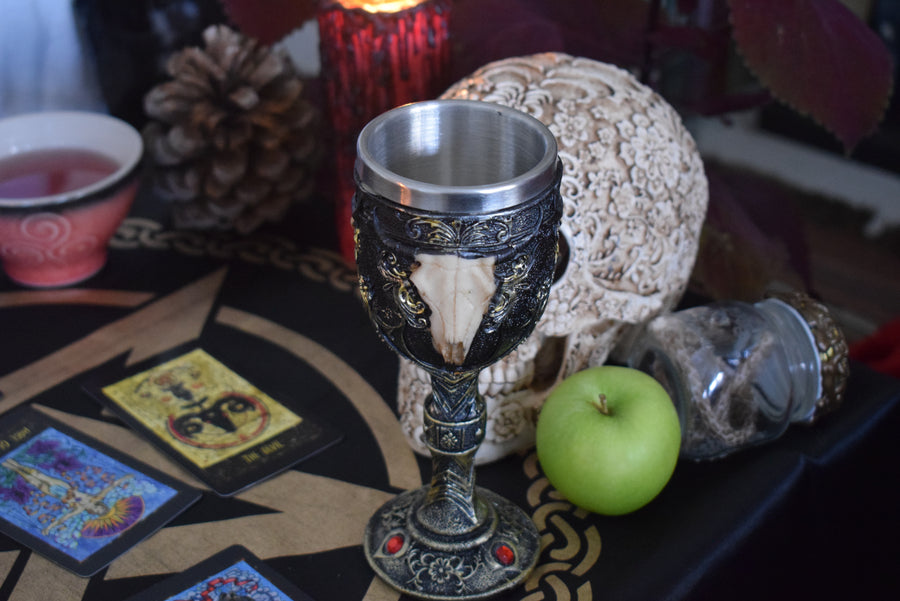 A goblet with a 3d cow skull with horns adorns the outside as it sits on an altar with a skull, snake skin, tarot cards and candle in the background