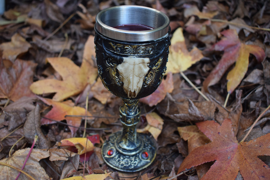 A goblet with a 3d cow skull with horns adorns the outside as it sits filled with wine on a bed of autumn leaves