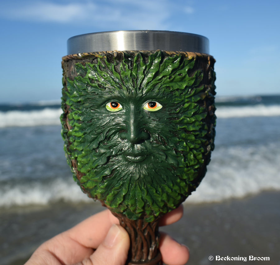 A hand holding a goblet depicting the green man with the ocean in the background.