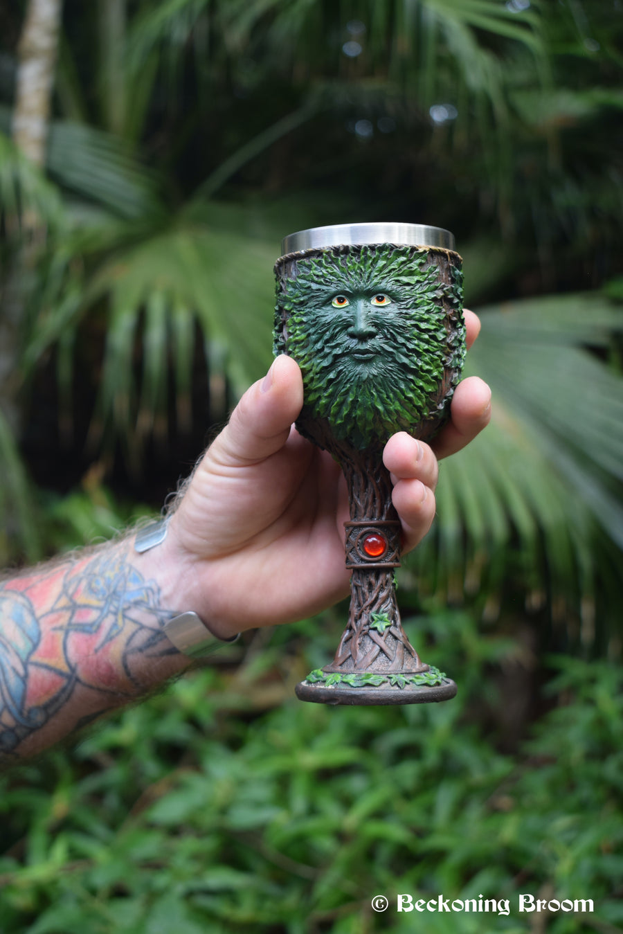 A hand holds a goblet depicting the green man with greenery in the background.
