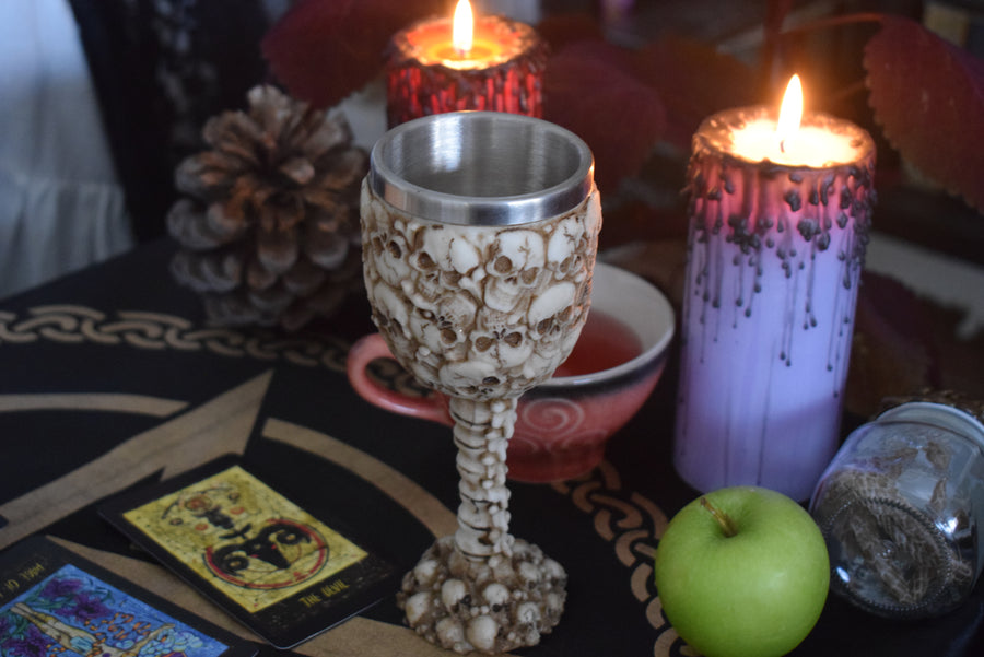 A goblet with 3d skulls all over it sits on an altar with candles, tarot cards, an apple and a jar of snake skin
