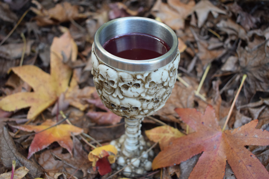A goblet with 3d skulls all over it sits filled with wine on a bed of autumn leaves