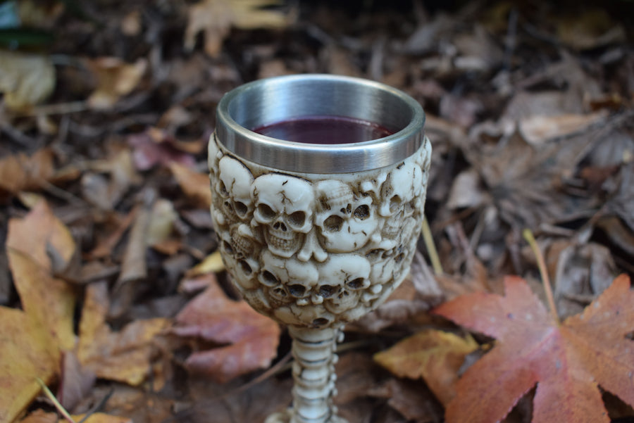 A goblet with 3d skulls all over it sits filled with wine on a bed of autumn leaves