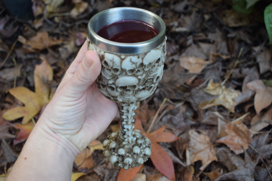 A hand holds a goblet with 3d skulls all over it sits filled with wine on a bed of autumn leaves